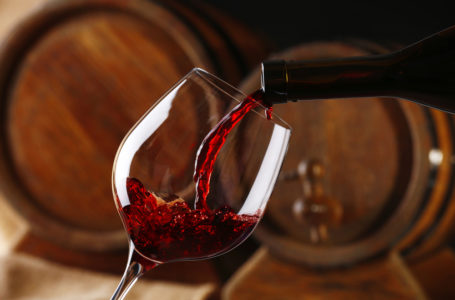 Pouring red wine from bottle into glass with wooden wine casks on background